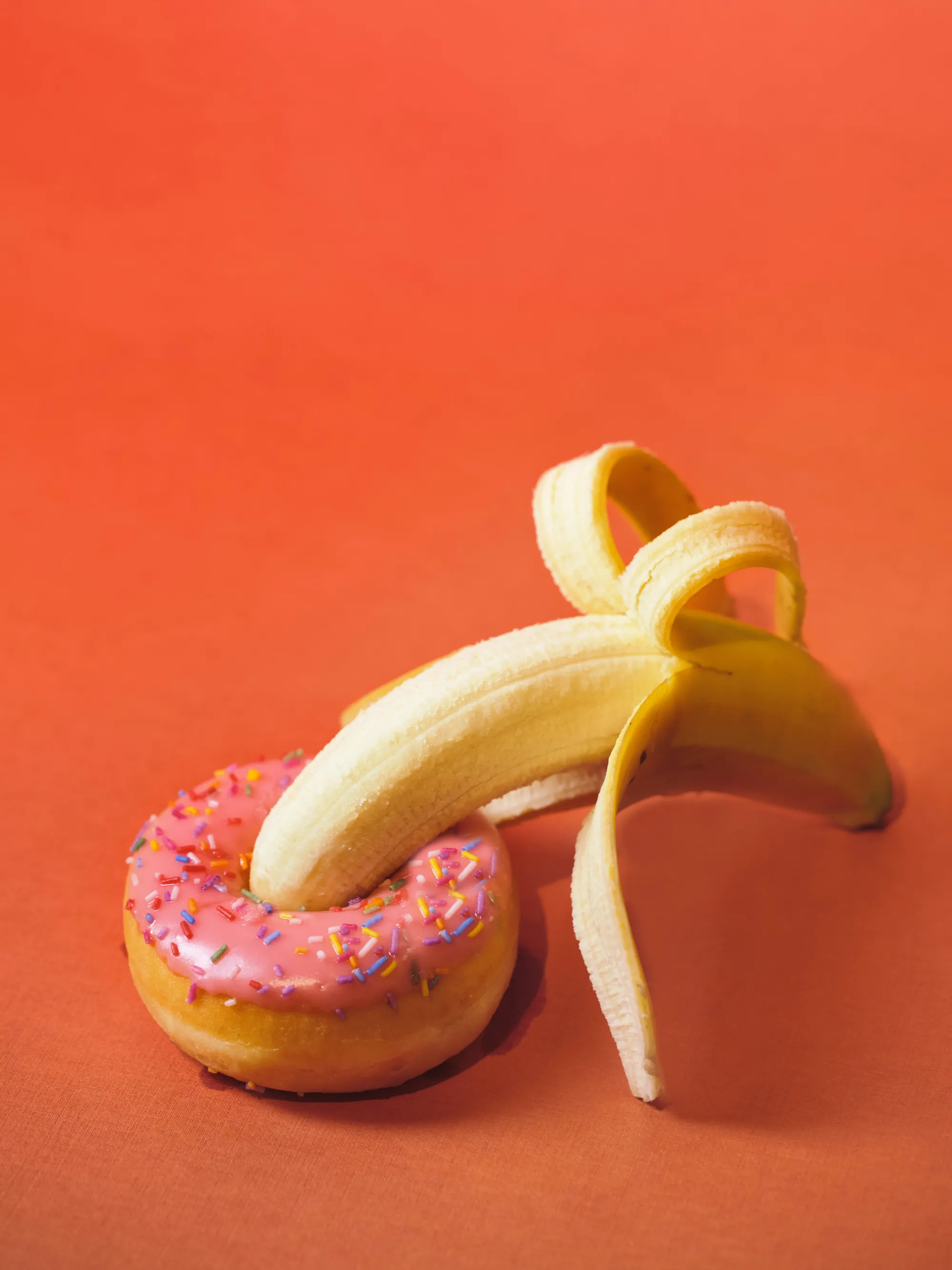 Un banano pelado atravesando una dona rosada con chispas